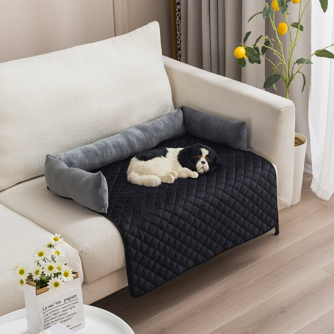 Dog lying on a black pet bed with gray pillows on a beige couch in a living room.